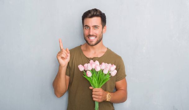 Guapo joven sobre gris pared grunge sosteniendo flores ramo muy feliz señalando con la mano y el dedo a un lado
 - Foto, Imagen