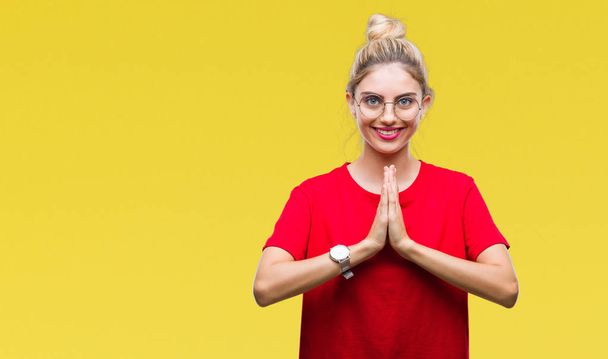 Young beautiful blonde woman wearing red t-shirt and glasses over isolated background praying with hands together asking for forgiveness smiling confident. - Photo, Image