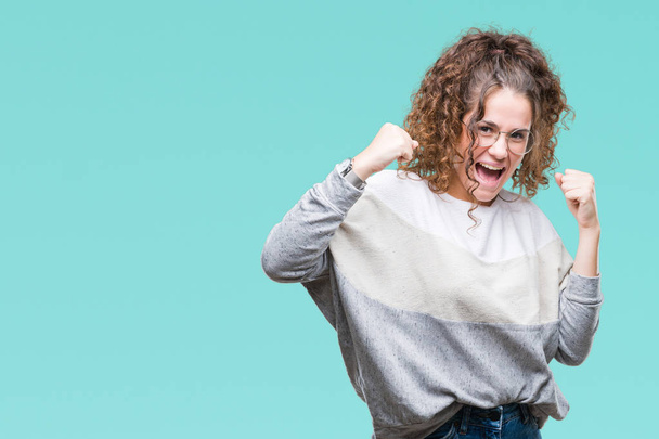 Beautiful brunette curly hair young girl wearing glasses over isolated background very happy and excited doing winner gesture with arms raised, smiling and screaming for success. Celebration concept. - Фото, изображение