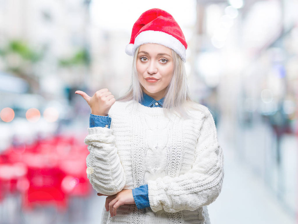 Νεαρή ξανθιά γυναίκα που φοράει καπέλο Χριστουγέννων πέρα από το απομονωμένο υπόβαθρο χαμογελώντας με χαρούμενο πρόσωπο αναζητούν και τοποθετώντας το δείκτη στην πλευρά με τον αντίχειρα. - Φωτογραφία, εικόνα