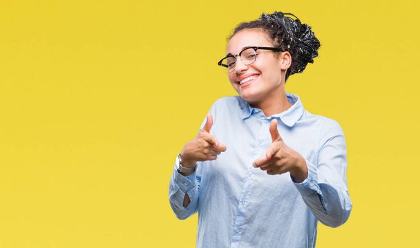 Joven chica de negocios afroamericana trenzado pelo usando gafas sobre fondo aislado señalando los dedos a la cámara con la cara feliz y divertida. Buena energía y vibraciones
. - Foto, Imagen