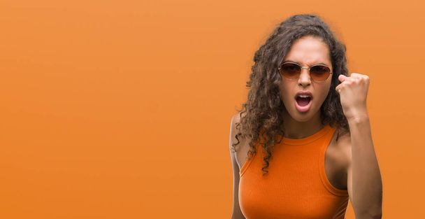 Молодая латиноамериканка в летнем наряде раздражена и разочарована криками гнева, безумна и криками с поднятой рукой, концепция гнева
 - Фото, изображение