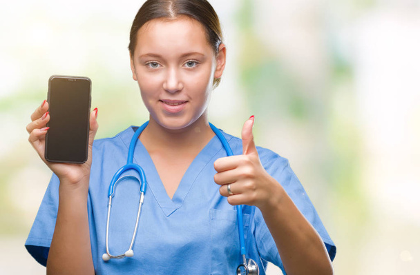 Young caucasian doctor woman showing smarpthone over isolated background happy with big smile doing ok sign, thumb up with fingers, excellent sign - Foto, afbeelding