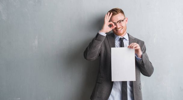 Young redhead  business man over grey grunge wall holding blank paper contract with happy face smiling doing ok sign with hand on eye looking through fingers - Photo, Image