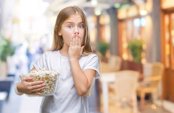 junge schöne Mädchen essen Popcorn-Snack isoliert Hintergrund Deckel Mund mit Hand schockiert vor Scham für Fehler, Ausdruck der Angst, Angst in der Stille, geheimes Konzept - Foto, Bild