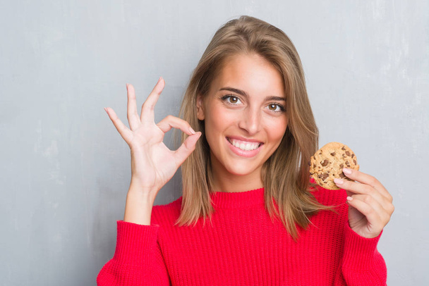 Красива молода жінка над гранжевою сірою стіною їсть шоколадний чіп з кулінарним знаком з пальцями, відмінний символ
 - Фото, зображення