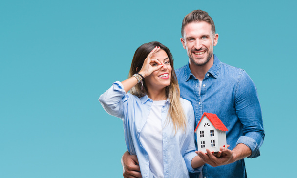 Young couple in love holding house over isolated background with happy face smiling doing ok sign with hand on eye looking through fingers - Фото, изображение