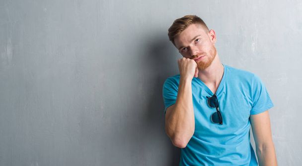 Молодой рыжеволосый мужчина на серой гранж-стене в повседневной одежде думает устало и скучно с депрессией проблемы со скрещенными руками
. - Фото, изображение