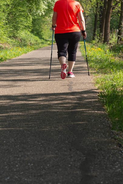 lady nordic walking in springtime park with red jacket and bright sun shining for happiness exercise - Foto, afbeelding