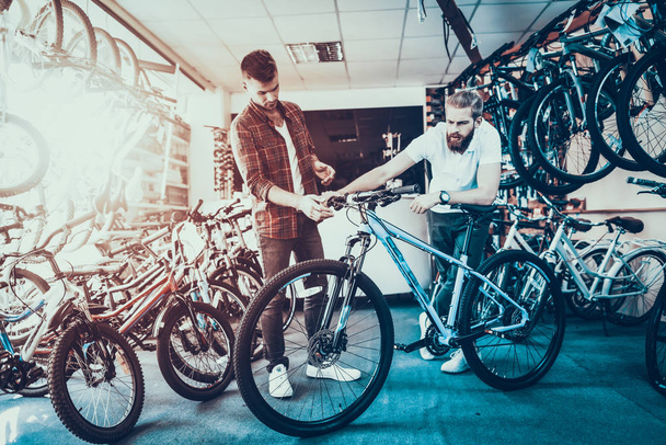 Consultant Shows Bicycle to Client in Sport Shop. Portrait of Young Male Shop Assistant Wearing White T-Shirt Helps in Bike Choosing. Happy Seller in Sport Store with Row of Bikes on Backround - Foto, afbeelding