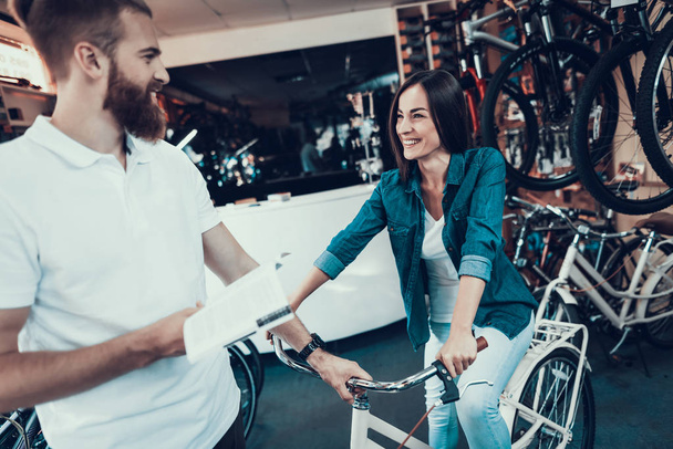 Girl Chooses City Bike with Seller in Sport Store. Handsome Bearded Shop Assistant Helps Young Client with White Vintage Cycle and Holds Instruction in Hands. Woman Try Riding Indoors. - Foto, Imagem