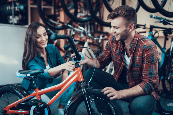 Happy Smiling Couple Chooses Kids Bicycle in Store. In Sport Cycle Workshop Young Beautiful Girl In Denim Clothes Talking with Handsome Consultant Wearing Checkered Shirt and Laughs - Фото, изображение