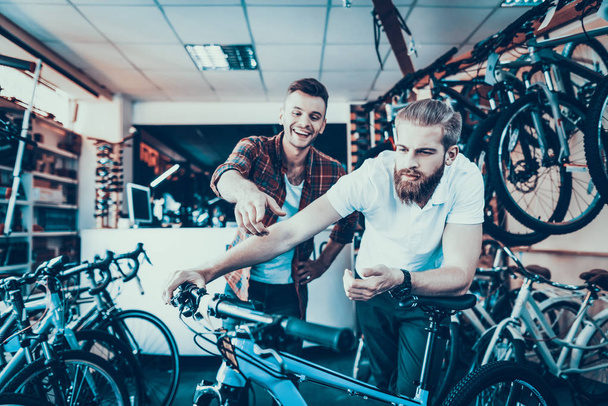 Consultant Shows Bicycle to Client in Sport Shop. Portrait of Young Shop Assistant Wearing White T-Shirt Helps in Mountain Bike Choosing. Happy Seller in Sport Store with Row of Bikes on Backround - Photo, Image
