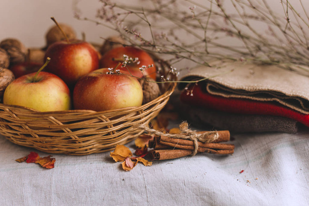 Still life with apples in a wicker basket, cloth, walnuts, flower petals and cinnamon sticks wrapped in twine. Concept of fall or winter home comfort. - Photo, Image