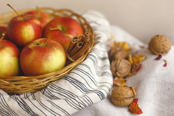 Still life with apples in a wicker basket stand on a striped linen fabric, walnuts, flower petals and cinnamon sticks wrapped in twine. Thanksgiving card creation for fall and autumn concepts. - Фото, зображення