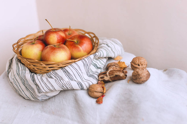 Still life with apples in a wicker basket stand on a striped linen fabric, walnuts, flower petals and cinnamon sticks wrapped in twine. Thanksgiving card creation for fall and autumn concepts. - Photo, Image