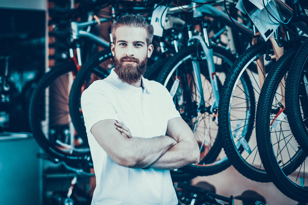 Salesman with Crossed Hands Poses in Bicycle Shop. Portrait of Cheerful Smiling Bearded Caucasian Man Wearing White T-Shirt Looking at Camera Standing Near Bicycles in Workshop - Zdjęcie, obraz