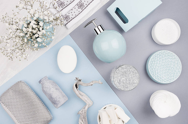 Fashion cosmetic products set on pastel blue, grey color paper - white soap, towel, flowers, soap dispenser, blue ceramic vase, silver cosmetic bag, top view. - Фото, изображение