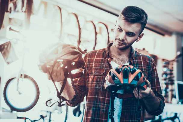 Guy Looks Closely at Helmets for Bicycle Rides. Portrait of Caucasian Customer Wearing Red Checkered Shirt Chooses Between Different Modern Goods in Sport Bicycle Store with Blurred Backround - 写真・画像
