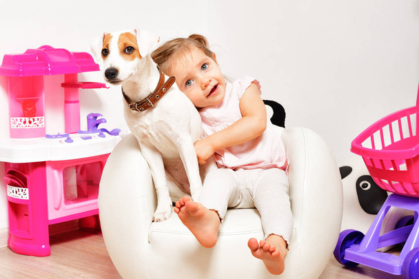 Petite fille mignonne étreignant son chien, assise sur une chaise dans la salle de jeux
 - Photo, image