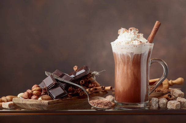 Hot chocolate with cream, cinnamon, chocolate pieces and various spices on a brown background. Copy space for your text. - Photo, Image