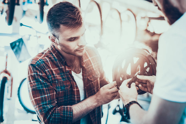 Man Looks at Helmet for Bike Rides in Sport Store. Young Caucasian Smiling Guy Wearing Red Checkered Shirt Buys Modern Helmet Talks with Bearded Shop Assistant in Bicycle Shop - Foto, Imagem