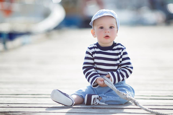 A kid, a boy with big brown eyes, a slightly open mouth, is dressed in a striped T-shirt, blue sneakers and blue jeans, a blue cap is on his head, crawls alone on a wooden pier, plays with a mooring rope, a portrait against the background of boats - Foto, Imagem
