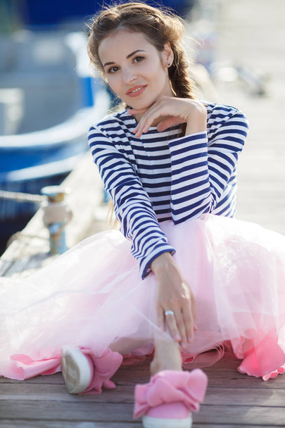 A beautiful young woman, a brunette with long hair plaited in a braid, dressed in a pink transparent skirt and a striped moth jersey, spends time sitting on a wooden pier by the sea next to the moored boat - Foto, Bild