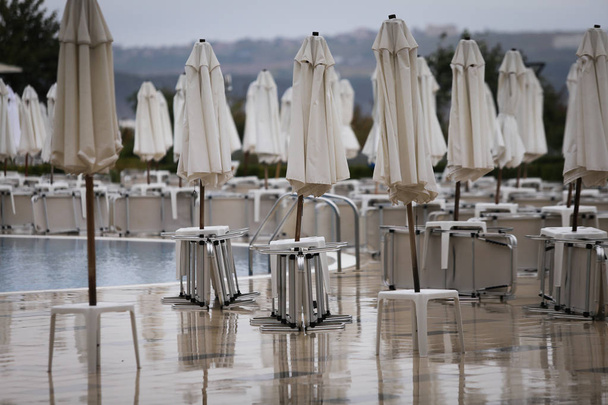 Sombrillas cerradas y sillas de césped alrededor de una piscina de hotel en temporada baja, sin personas en un día frío y nublado, a finales de otoño en la costa búlgara
 - Foto, imagen
