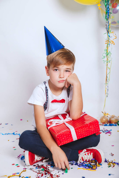 Cute sad little boy in birthday cap holding red present. Holidays concept. - Φωτογραφία, εικόνα