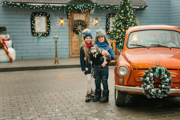 Little boy and girl with piggy standing near old car while snow falling - Photo, Image