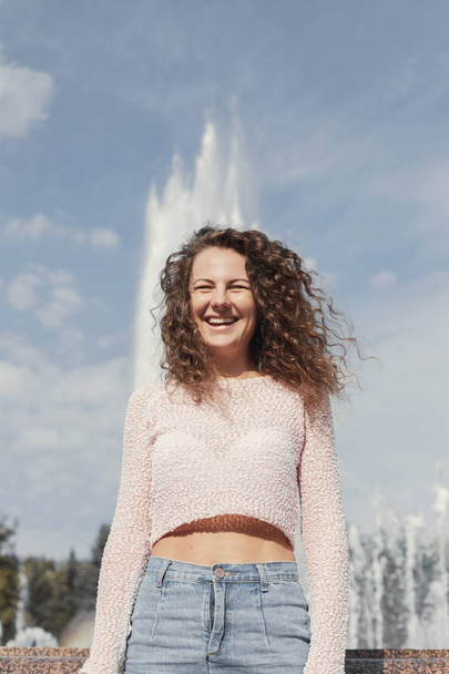 Наружный снимок горячей женщины пинап в модном розовом топе и джинсовой юбке, смеющиеся нагрузки, улыбаясь радостно позируя над парком с фонтаном фона, гуляя наслаждаясь теплым солнечным днем, фотографируя
. - Фото, изображение