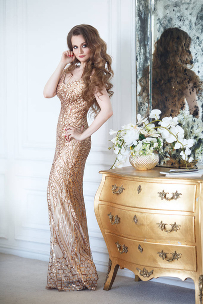 Slender elegant woman in an evening long gold dress in a chic interior. - Foto, afbeelding