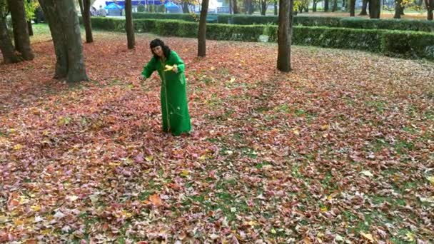 Mooie gelukkige vrouw in een groene jurk, verzamelt bladeren in de herfst park en gooit ze omhoog. Gouden herfst concept. Buitenshuis. - Video