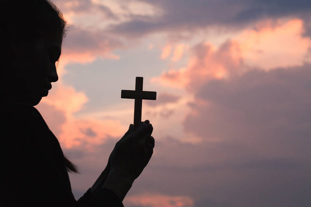 Silhouette della mano della donna che tiene il sollevamento santo della croce cristiana con lo sfondo leggero del tramonto
. - Foto, immagini