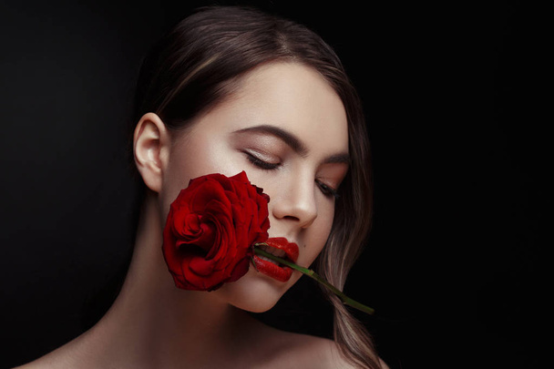 Ομορφιά πορτρέτο του ένα κομψό σέξι κορίτσι με ένα κόκκινο τριαντάφυλλο στα δόντια της πάνω σε μαύρο φόντο. - Φωτογραφία, εικόνα