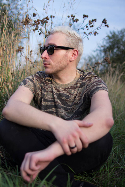 Po stronie portretu młodych atrakcyjnych Brodaty Blondynka facet sobie czarne okulary przeciwsłoneczne i siedzi na trawie pola na tle pola. - Zdjęcie, obraz