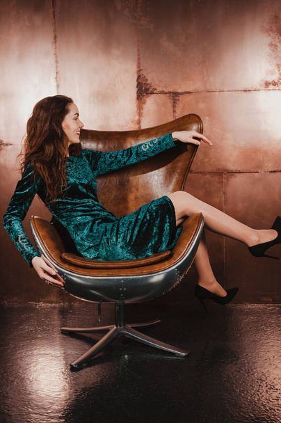 Jovem atraente em um vestido de veludo verde azul, sentado em uma cadeira marrom de couro, se divertindo, sorrindo. Cabelos longos, pernas, saltos altos
 - Foto, Imagem