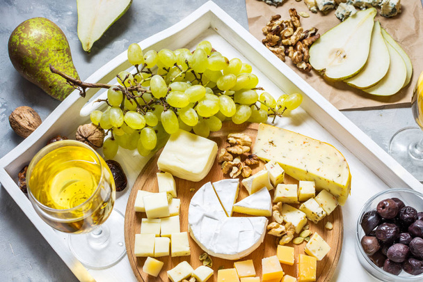 Различные виды сыра на деревянной доске, оливки, фрукты, миндаль и бокалы вина на белом подносе на бетонном столе
 - Фото, изображение