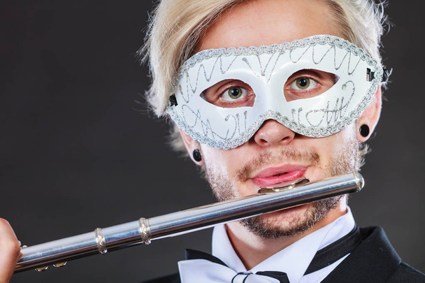 Holidays, people and celebration concept. Elegant young guy wearing suit white shirt bow tie and carnival venetian mask playing flute instrument on dark - Fotó, kép