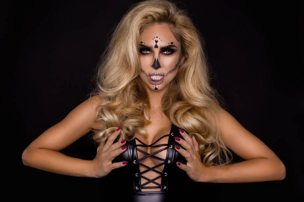 Mujer rubia sexy en maquillaje de Halloween y traje de cuero sobre un fondo negro en el estudio. Esqueleto, monstruo y bruja
. - Foto, imagen