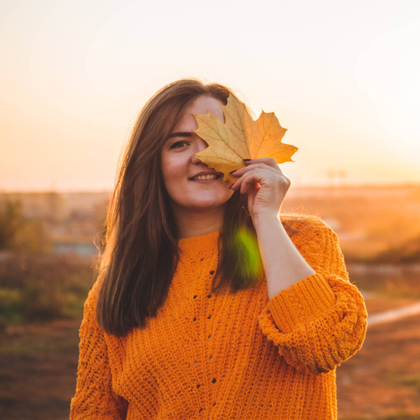 junge Frau in orangefarbenem Pullover mit gelben Blättern, Außenporträt im sanften, sonnigen Tageslicht. Herbst. Sonnenuntergang. gemütlich - Foto, Bild