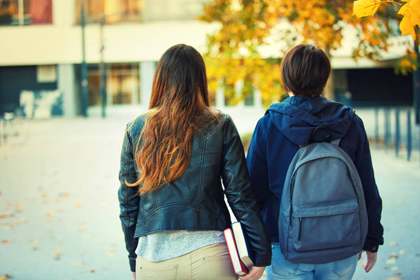 Rear view of two women students going to school or college carrying backpack and holding books in hand. Autumn outdoors friends walking educational concept.  - 写真・画像
