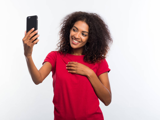 Smiling happy young african american girl in red t-shirt making selfie photo on smartphone over white background. Technology concept. - Foto, Imagem