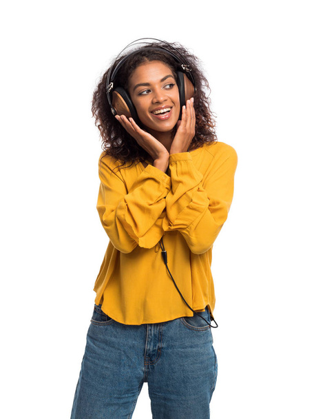 Atractiva joven afroamericana escuchando música con auriculares y bailando sobre fondo blanco. Chica con capucha amarilla. Grabado en estudio. Concepto musical
. - Foto, Imagen