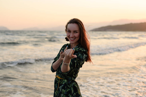 Πορτρέτο του μια νεαρή γυναίκα στην παραλία στο κόκκινο ηλιοβασίλεμα, νεύει με εσάς, καλώντας να έχουν τη διασκέδαση. - Φωτογραφία, εικόνα