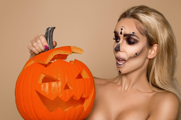 Σέξι ξανθιά γυναίκα στο μακιγιάζ Απόκριες κατέχουν μια κολοκύθα σε ένα φόντο beigek στο στούντιο. Σκελετός make-up, τέρας και η μάγισσα - Φωτογραφία, εικόνα