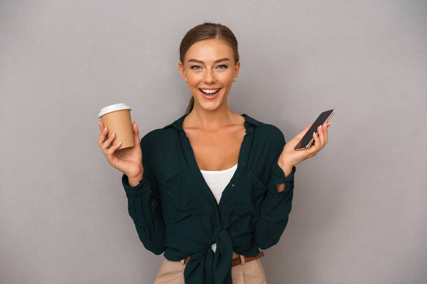 Image of excited beautiful business woman posing isolated over grey wall background drinking coffee using mobile phone. - Fotoğraf, Görsel