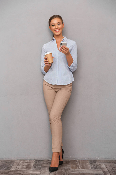 Image of happy business young woman posing isolated over grey wall background using mobile phone drinking coffee. - Valokuva, kuva