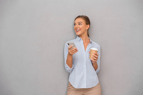 Image of happy business young woman posing isolated over grey wall background using mobile phone drinking coffee. - Foto, immagini